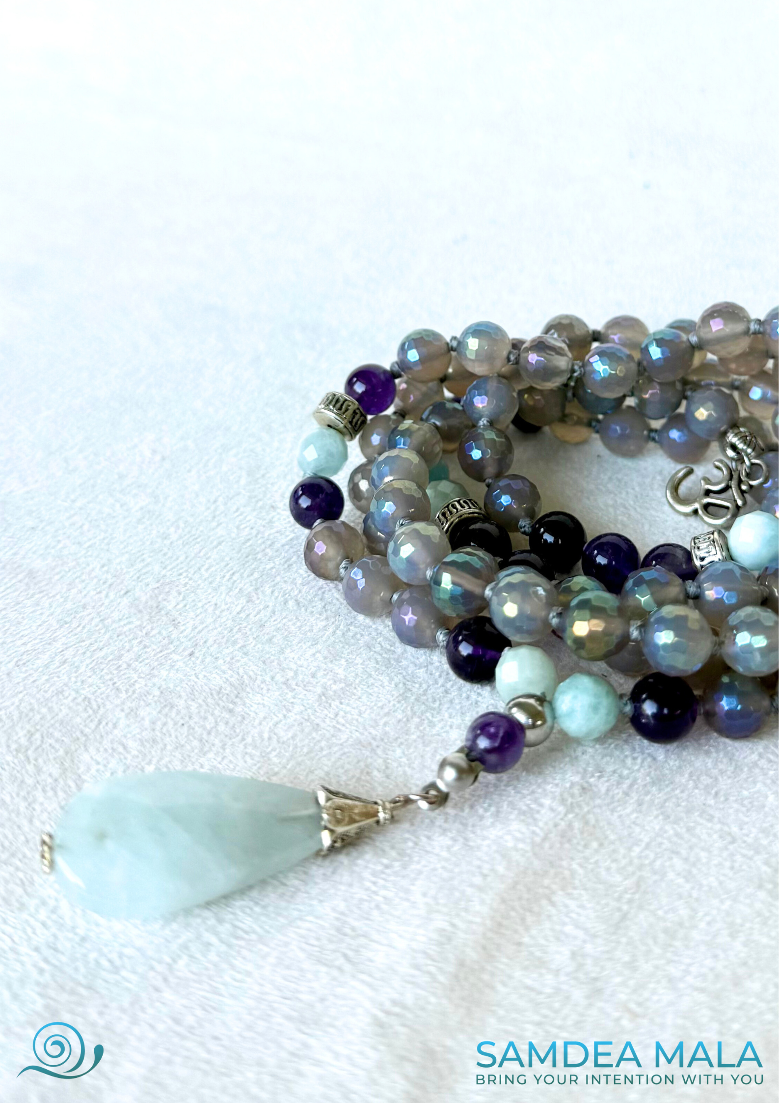 close-up of 108 spiritual beads and OM charm by Samdea Mala, with gray agate, amethyst, and aquamarine beads. Hand-knotted mala necklace with natural aquamarine pendant.