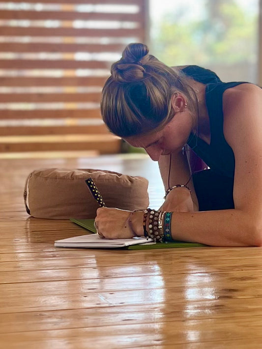 Samdea Mala A woman is writing on a white notepad. She is wearing a black tank top and several bracelets on her wrists. The notepad is on a wooden surface. The background is blurred but shows a wooden wall and a window with trees visible outside.