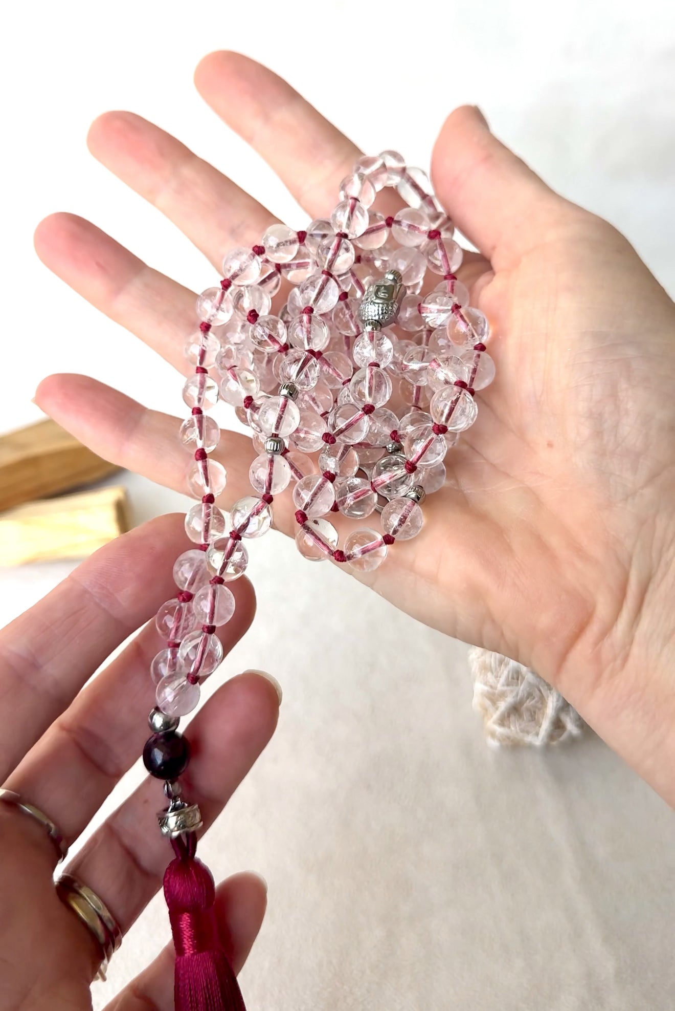 hands holding a clear quartz mala necklace and Buddha. Samdea Mala in clear quartz mala beads for meditation and mantra recitation On mani Pamde Hum. with garnet guru bead, and deep red thread and red tassel. Mala for meditation. 108 prayer beads, Spiritual gift.