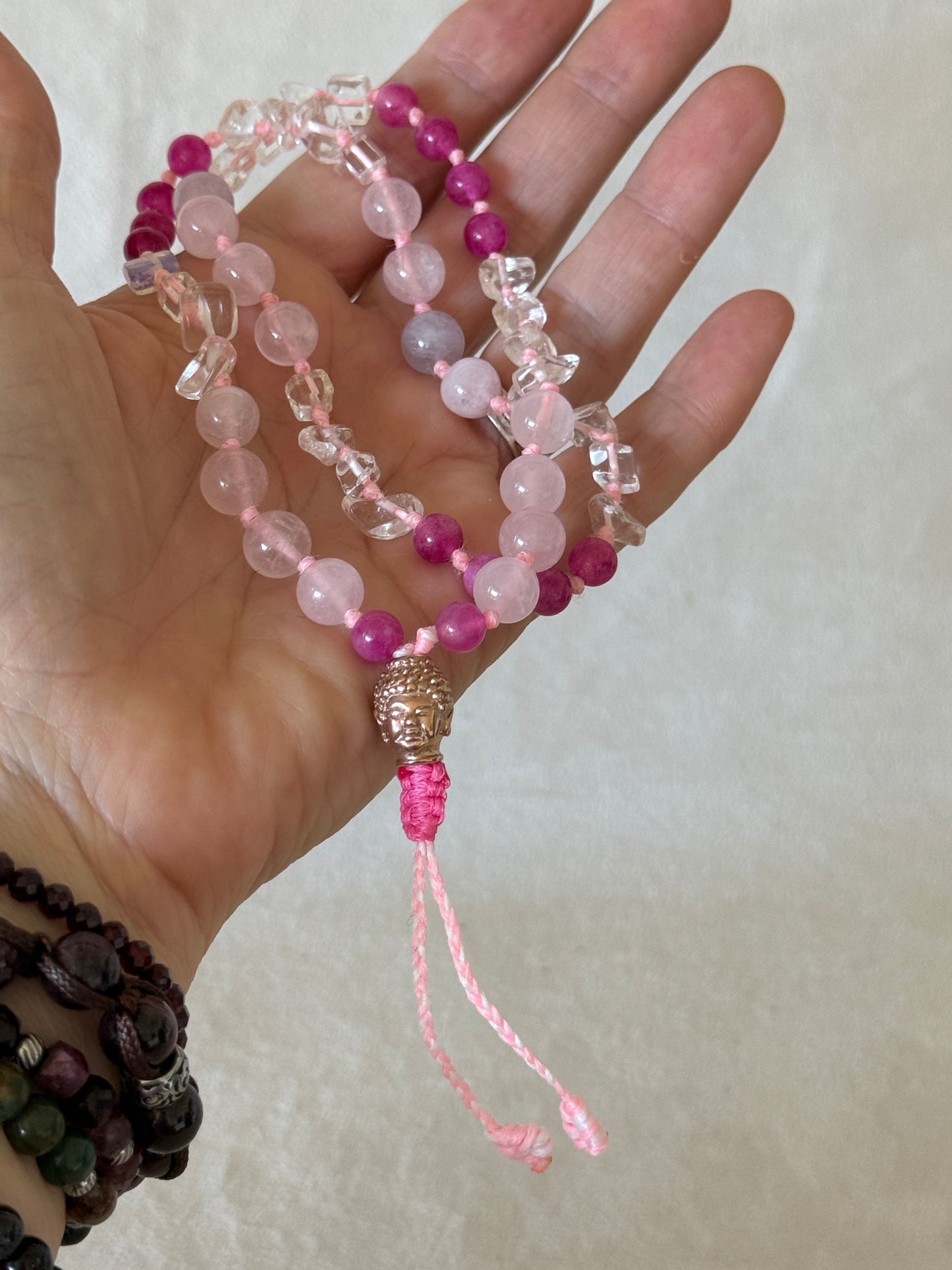 A hand holding a Handmade  Square Breathing Mala with pink agate, rose quartz, clear quartz, and a rose-gold-colored stainless steel Buddha head meru, designed for box breathing, meditation, and mindful pauses.