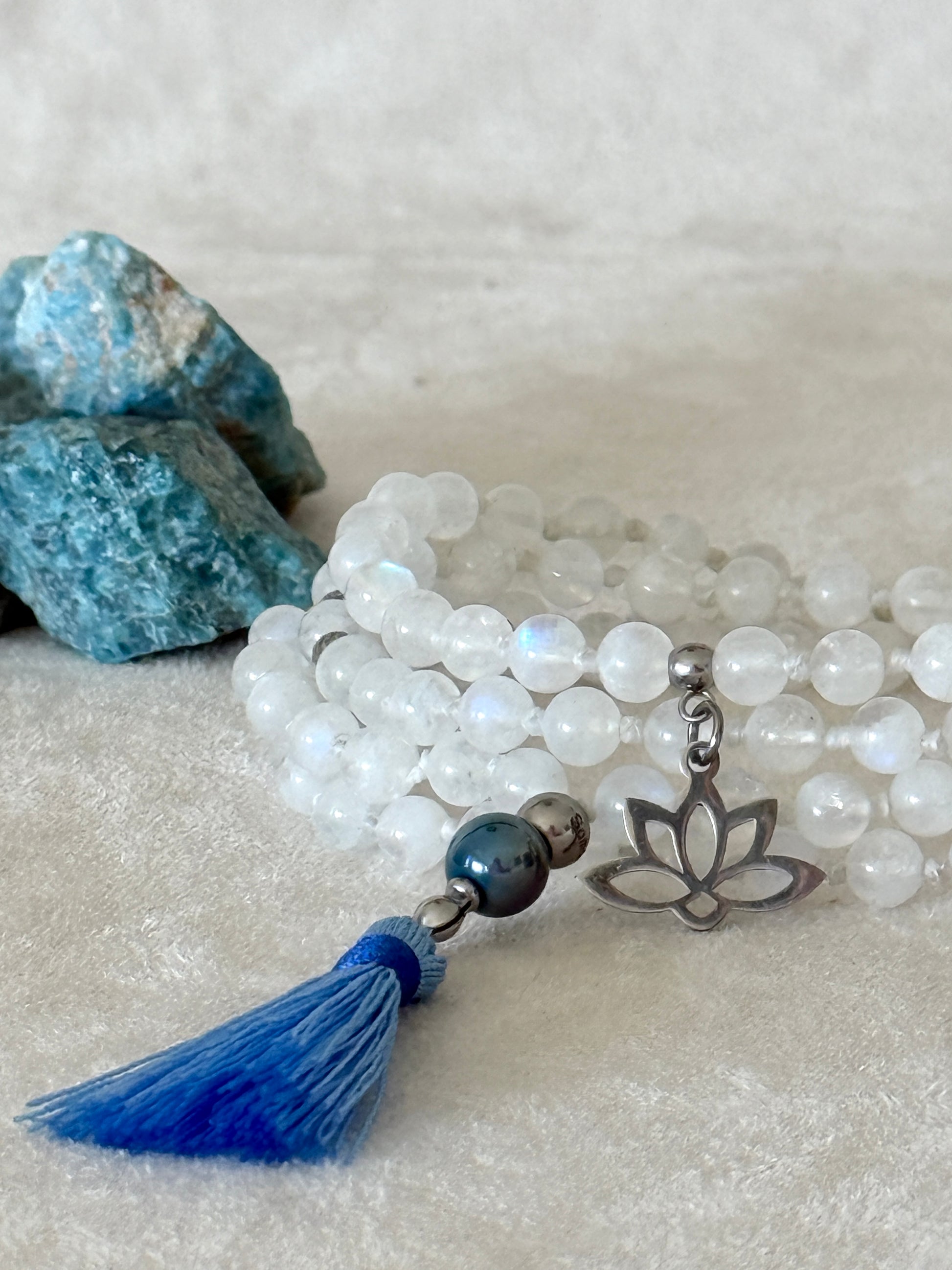 Close-up of Hand-knotted Rainbow Moonstone mala necklace with 108 prayer beads, 6 mm natural gemstone beads, Aqua Aura Quartz meru, stainless steel details, small lotus charm, and tassel, created for meditation, breathwork, mantra repetition, mindfulness, and intentional practice.