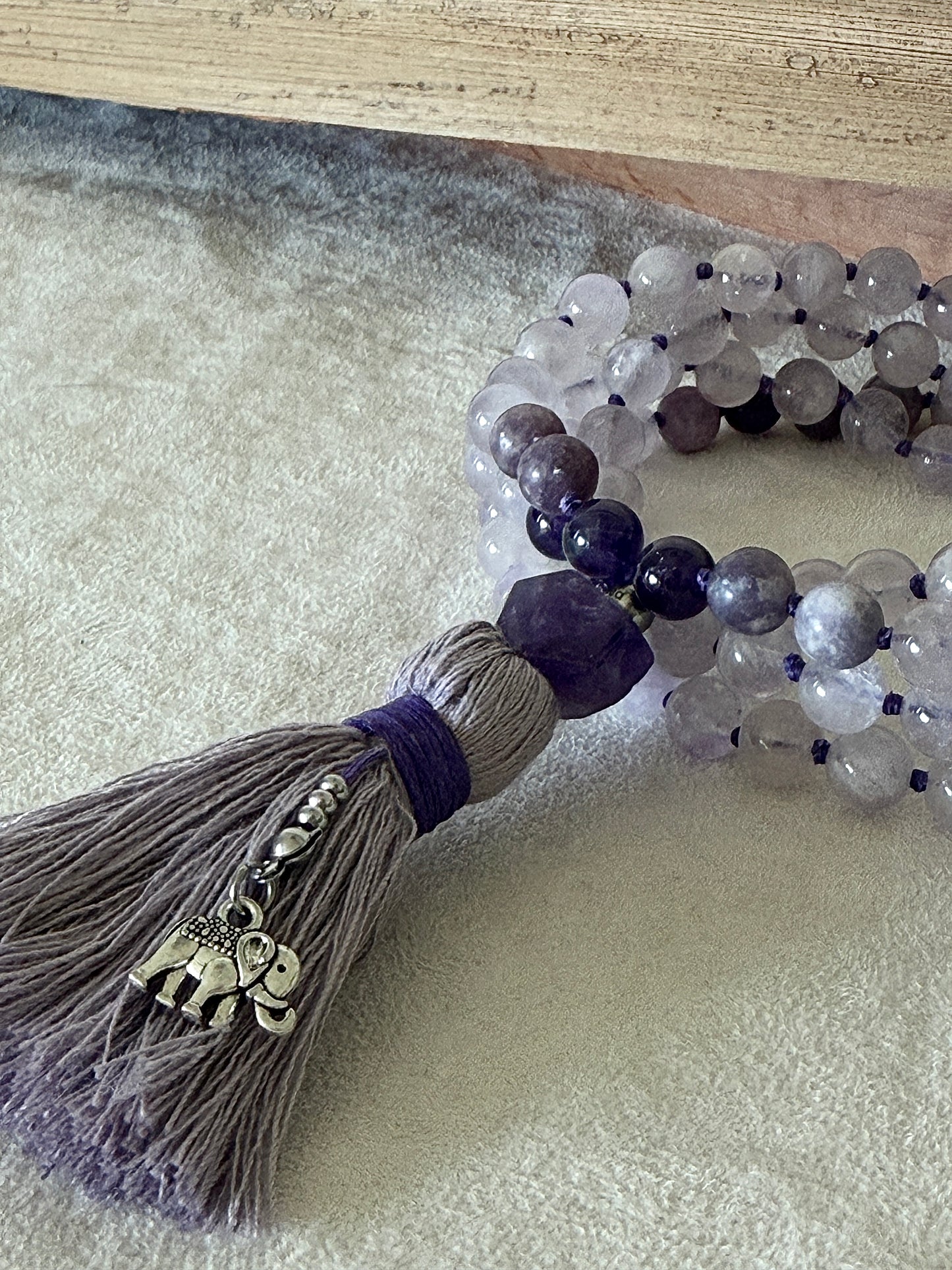 Close up of Purple mala necklace for meditation. Handmade 108 bead mala in netural gemstone 8 mm lavender jade, lepidolite and amethyst with cotton tassel and small elephant charm.