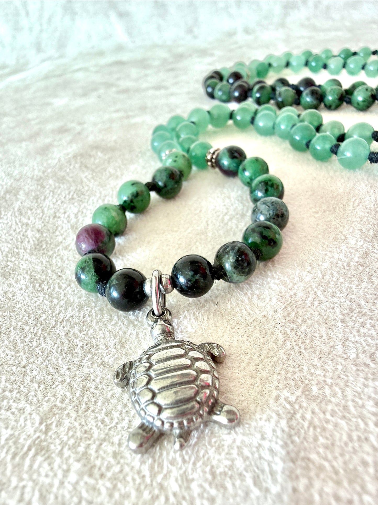 Close up of a  stainless steel turtle paendant Handmade Zoisite with Ruby and Aventurine greenmala bracelet with natural 6 mm gemstones, stainless steel turtle charm, adjustable macramé closure, and 108 bead mala necklace design for yoga, meditation, gentle growth, protection, and spiritual transformation. Samdea Mala