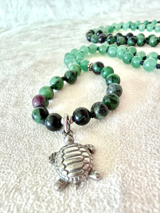 Close up of a  stainless steel turtle paendant Handmade Zoisite with Ruby and Aventurine greenmala bracelet with natural 6 mm gemstones, stainless steel turtle charm, adjustable macramé closure, and 108 bead mala necklace design for yoga, meditation, gentle growth, protection, and spiritual transformation. Samdea Mala