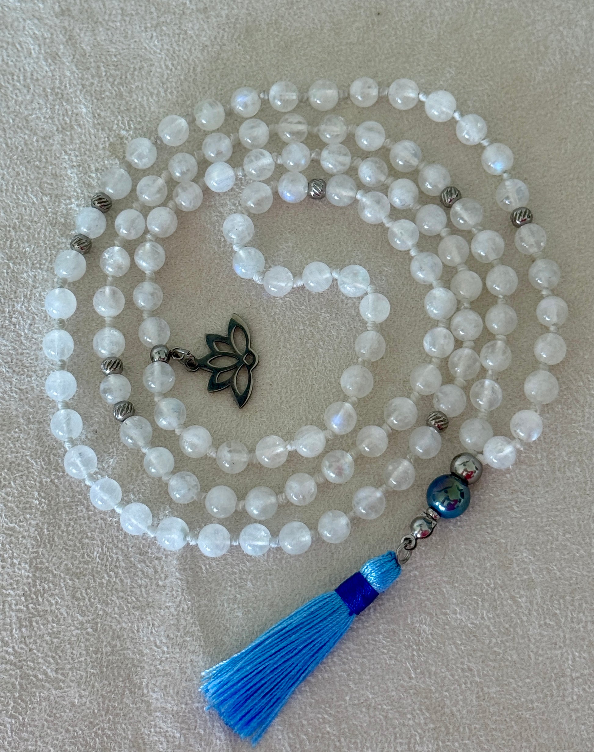 Full view of Hand-knotted Rainbow Moonstone mala necklace with 108 prayer beads, 6 mm natural gemstone beads, Aqua Aura Quartz meru, stainless steel details, small lotus charm, and tassel, created for meditation, breathwork, mantra repetition, mindfulness, and intentional practice.