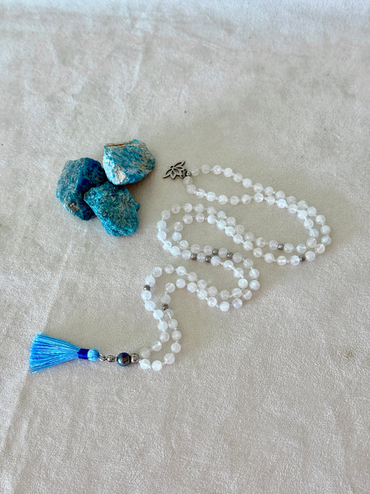 Full view of Hand-knotted Rainbow Moonstone mala necklace with 108 prayer beads, 6 mm natural gemstone beads, Aqua Aura Quartz meru, stainless steel details, small lotus charm, and tassel, created for meditation, breathwork, mantra repetition, mindfulness, and intentional practice.