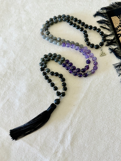 Shadow Work Mala with 108 natural gemstone beads in obsidian, amethyst, dark labradorite and lava stone, finished with a black tassel, Triquetra charm and stainless steel Om Mani Padme Hum mantra bead. Designed as a tactile support for meditation, reflection and shadow work.