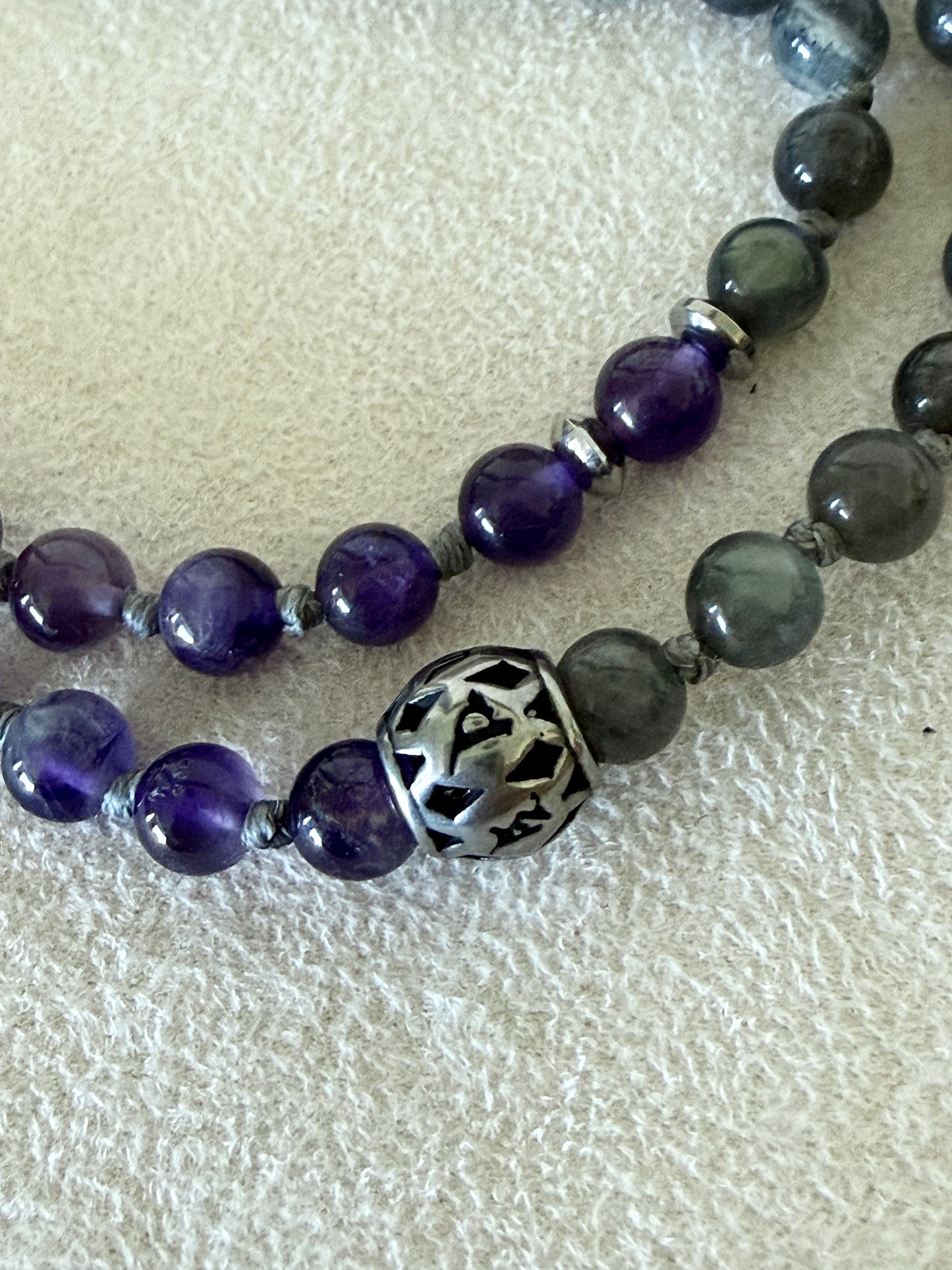 Close-up of the engraved steel Om Mani Padme Hum mantra bead on the Shadow Work Mini Mala, framed by amethyst and labradorite beads.