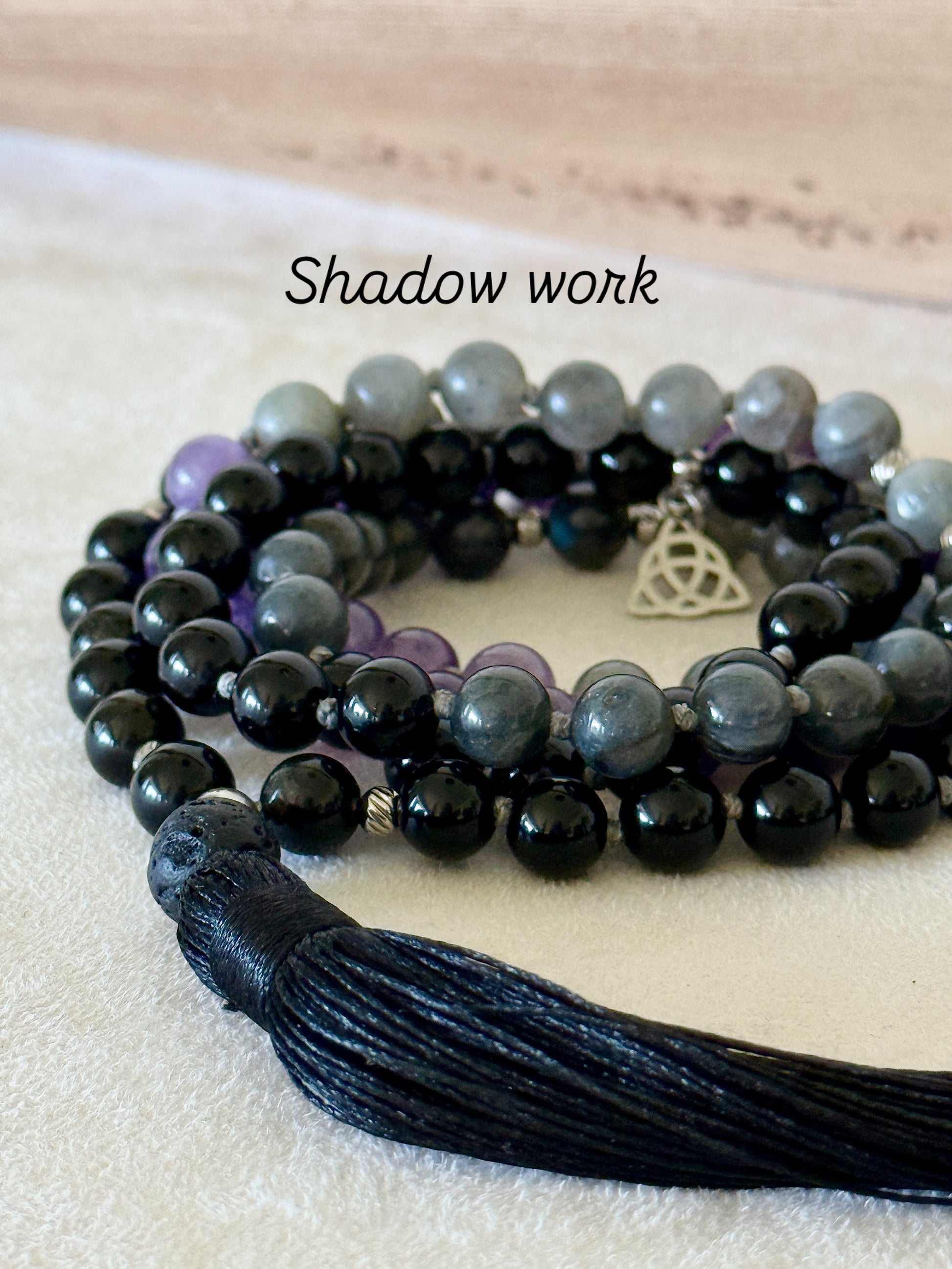 Shadow Work Mala with 108 natural gemstone beads in obsidian, amethyst, dark labradorite and lava stone, finished with a black tassel, Triquetra charm and stainless steel Om Mani Padme Hum mantra bead. Designed as a tactile support for meditation, reflection and shadow work.