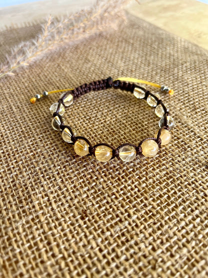 Shamballa handmade bracelet features light yellow citrine quartz gemstone beads on a brown cord. The bracelet has a sliding closure with yellow cord ends and small metal accents. The bracelet is displayed on a burlap background. 