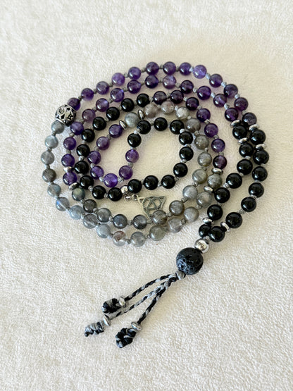 Top view of the coiled Shadow Work Mini Mala showing the full bead sequence, engraved silver guru bead, Triquetra charm, and tassel.
