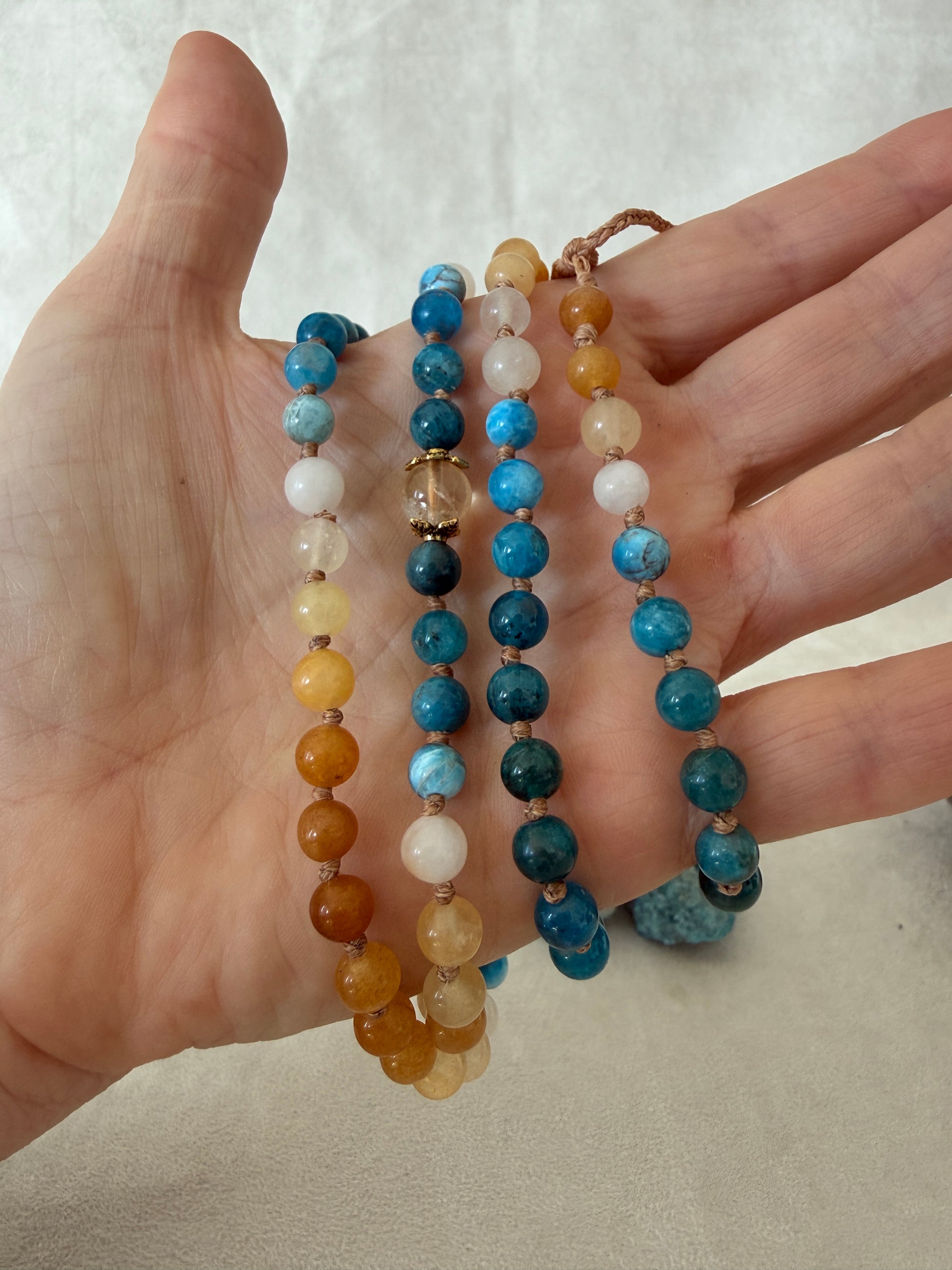 a Hand holding The Sun and the sea natural gemstone mala bracelet. Handmade Apatite and Yellow Jade mala bracelet with natural 6 mm gemstones, Citrine guru bead, adjustable macramé closure, and stainless steel details; a 108 bead mala necklace inspired by sea and sun, water and fire, for meditation, yoga, and intention practice.
