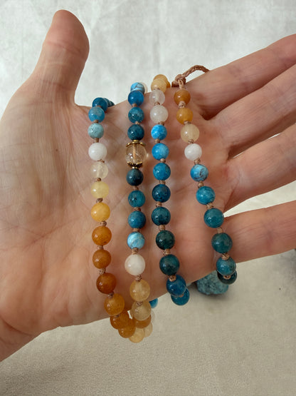 a Hand holding The Sun and the sea natural gemstone mala bracelet. Handmade Apatite and Yellow Jade mala bracelet with natural 6 mm gemstones, Citrine guru bead, adjustable macramé closure, and stainless steel details; a 108 bead mala necklace inspired by sea and sun, water and fire, for meditation, yoga, and intention practice.