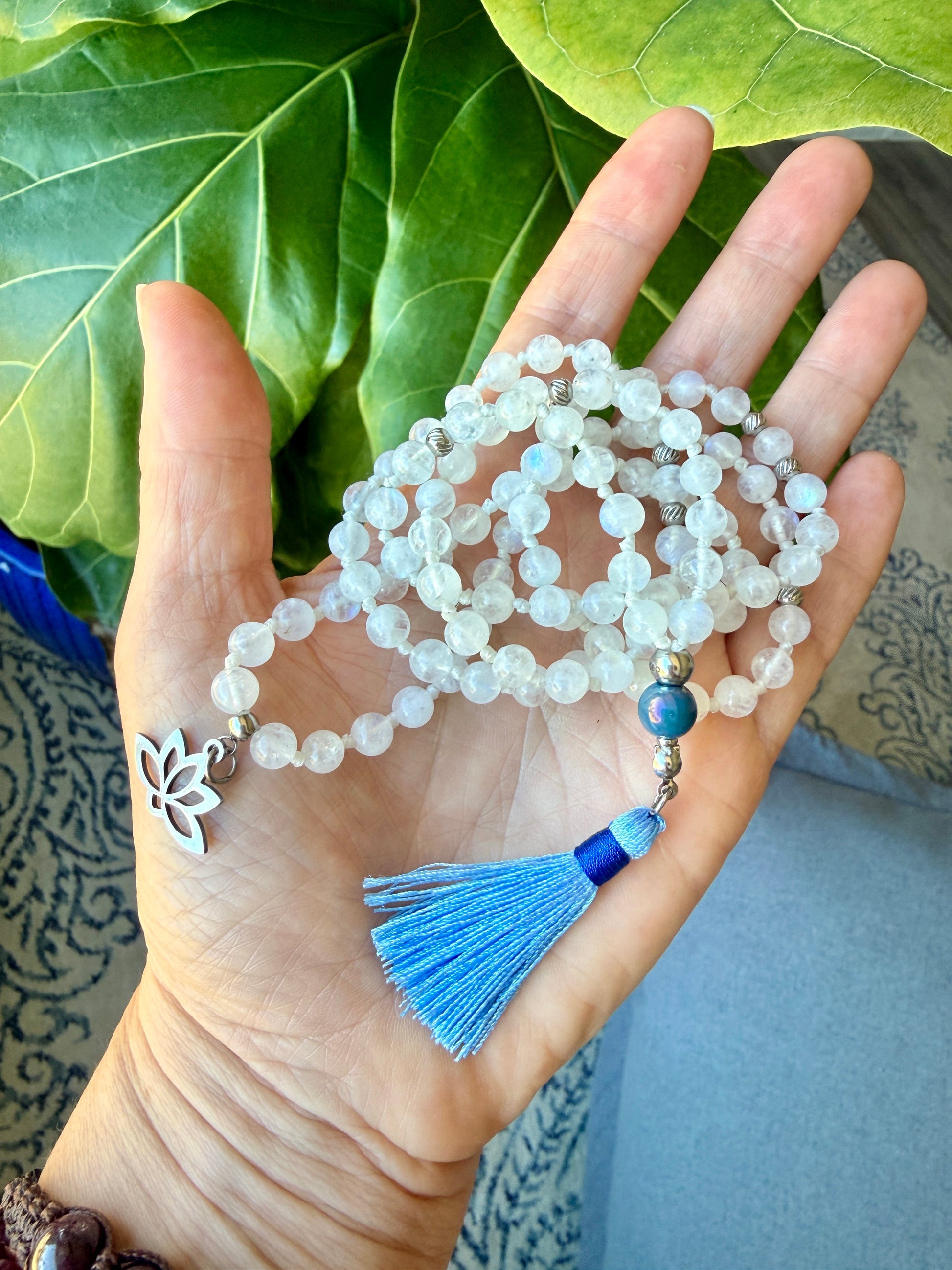 a hand holding a Handmade Rainbow Moonstone mala necklace with 108 prayer beads, 6 mm natural gemstone beads, Aqua Aura Quartz meru, stainless steel details, small lotus charm, and tassel, created for meditation, breathwork, mantra repetition, mindfulness, and intentional practice.