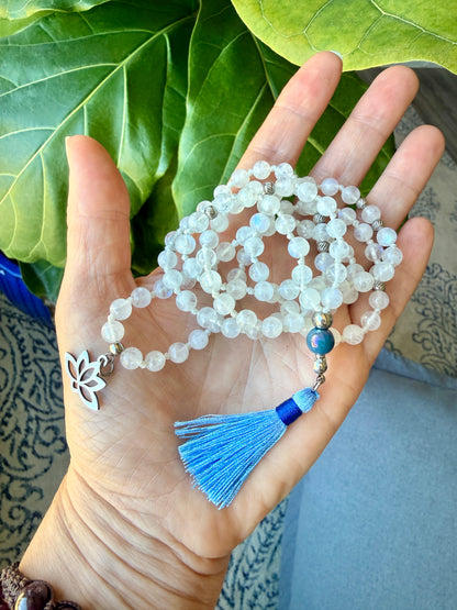 a hand holding a Handmade Rainbow Moonstone mala necklace with 108 prayer beads, 6 mm natural gemstone beads, Aqua Aura Quartz meru, stainless steel details, small lotus charm, and tassel, created for meditation, breathwork, mantra repetition, mindfulness, and intentional practice.